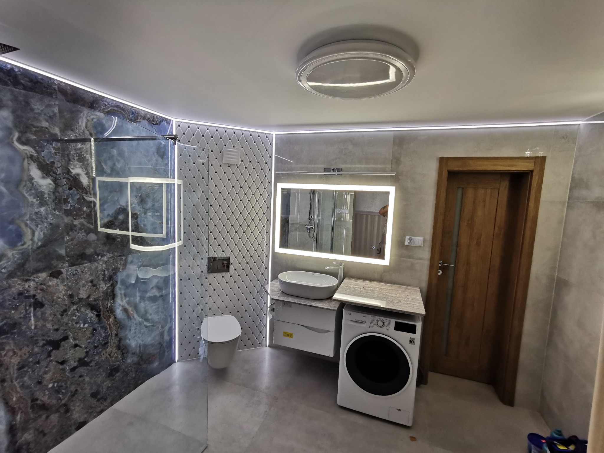 Nowoczesna łazienka z kabiną prysznicową z marmurową ścianą, podświetlanym lustrem nad umywalką nablatową, pralką obok szafki umywalkowej i podwieszaną toaletą na ścianie z mozaiką romboidalną...