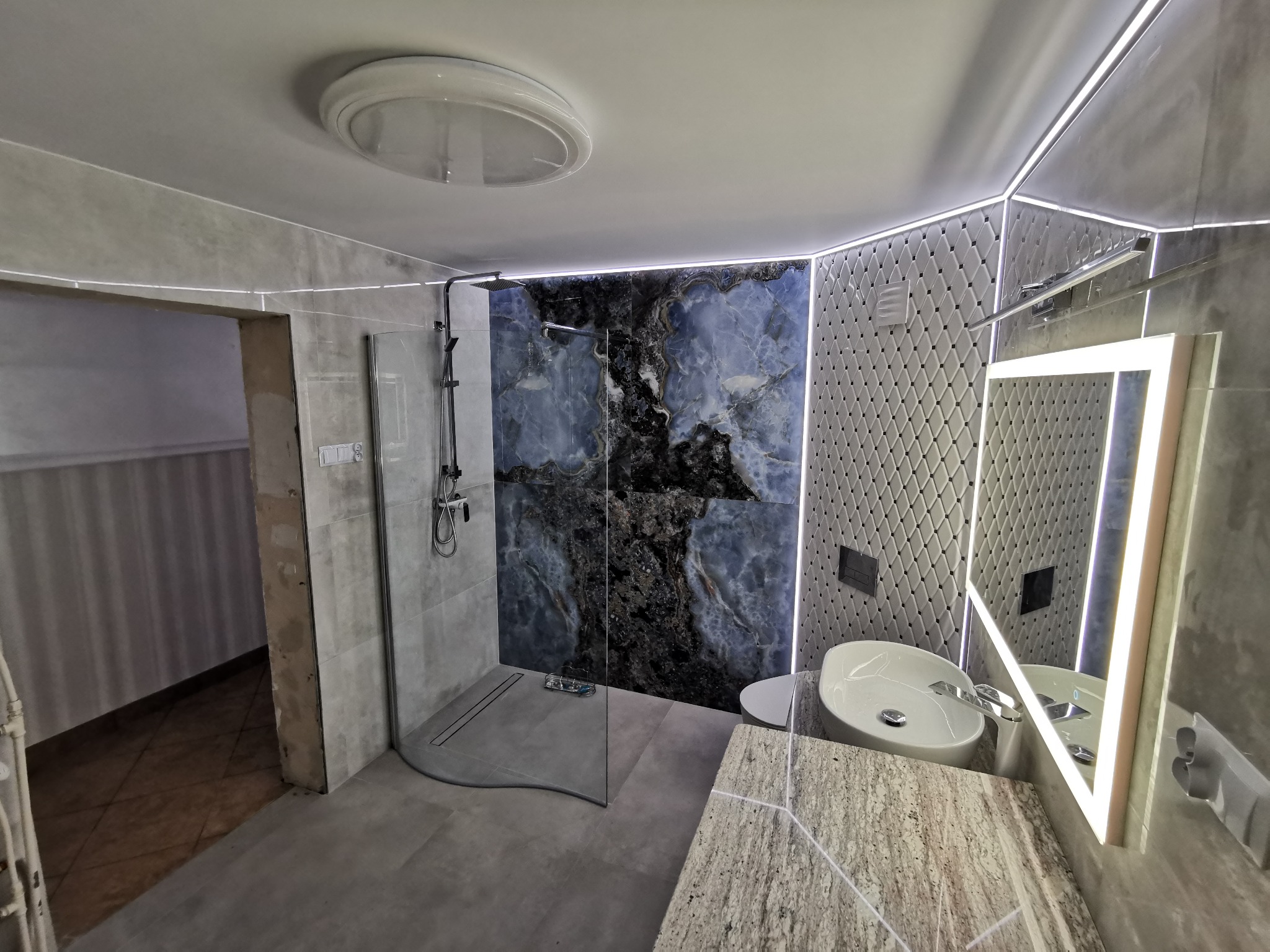 Nowoczesna łazienka po remoncie: kabina prysznicowa typu walk-in z czarną armaturą, ściana wyłożona płytkami imitującymi marmur w odcieniach granatu i szarości, umywalka nablatowa z podświetlanym...