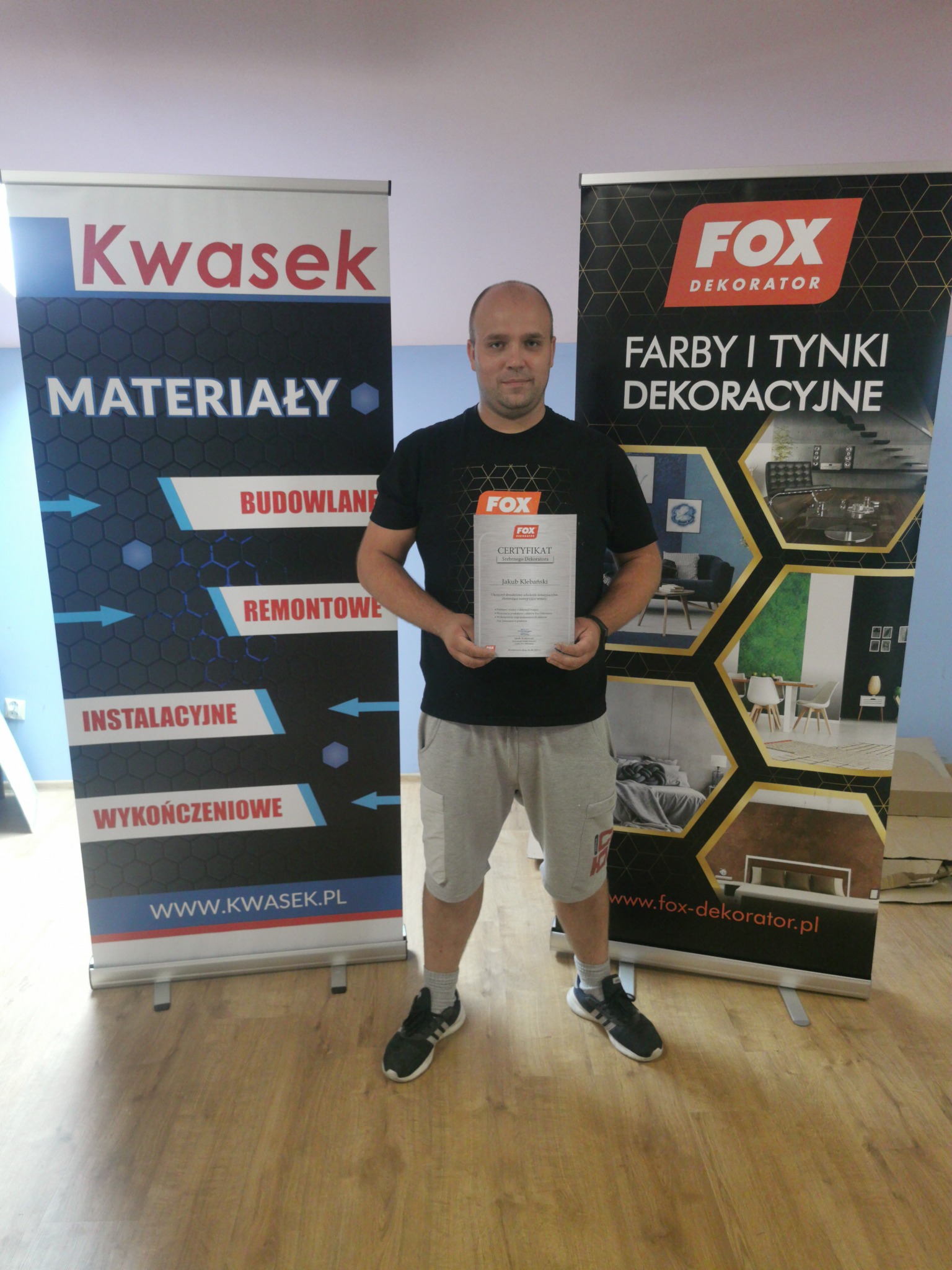 Mężczyzna trzyma certyfikat srebrnego dekoratora Fox, stojąc pomiędzy roll-upami reklamowymi firm Kwasek i Fox Dekorator prezentującymi materiały budowlane, remontowe, wykończeniowe oraz farby...