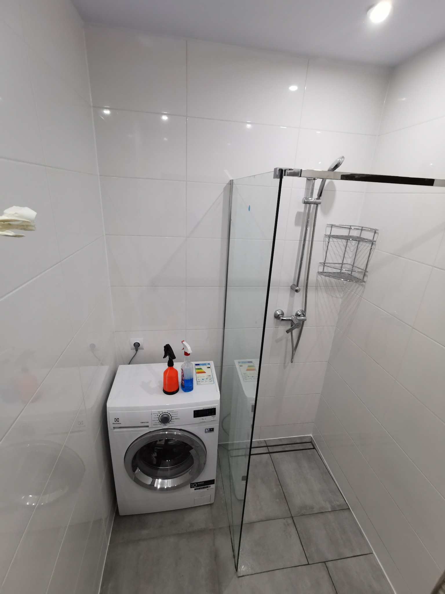 Jasna łazienka po remoncie z kabiną prysznicową typu walk-in, płytkami w odcieniach bieli i szarości oraz pralką Electrolux z butelkami środków czyszczących na górze.