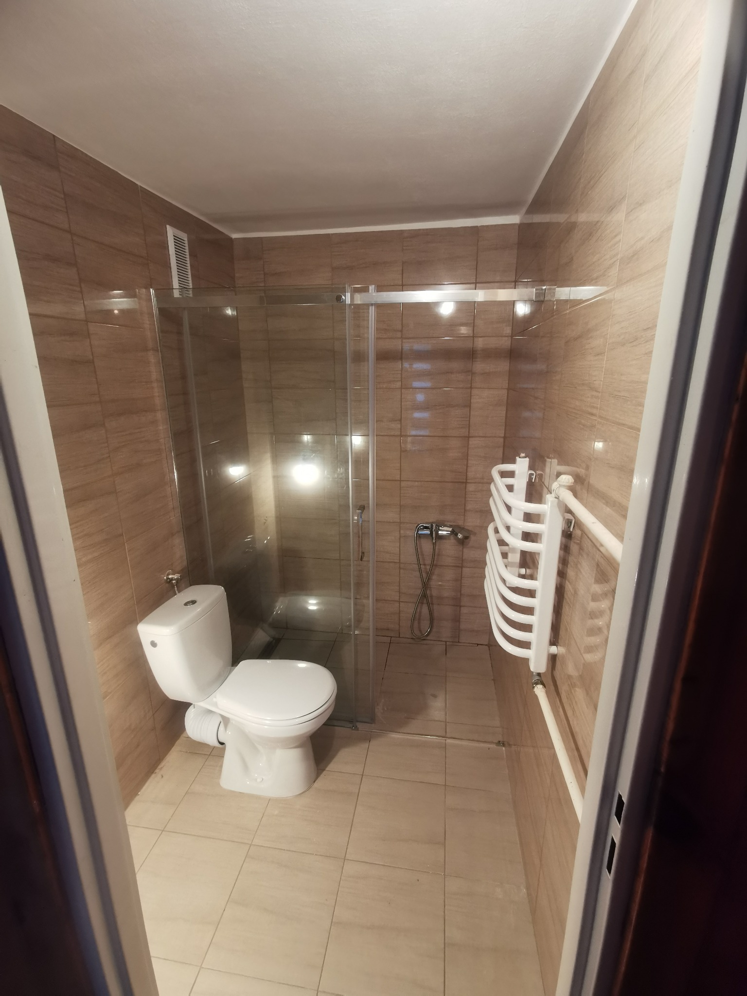 Wykończona łazienka z kabiną prysznicową z przesuwnymi drzwiami, wiszącym grzejnikiem drabinkowym i toaletą. Płytki w odcieniach beżu na ścianach i podłodze. Widok zza futryny drzwi.