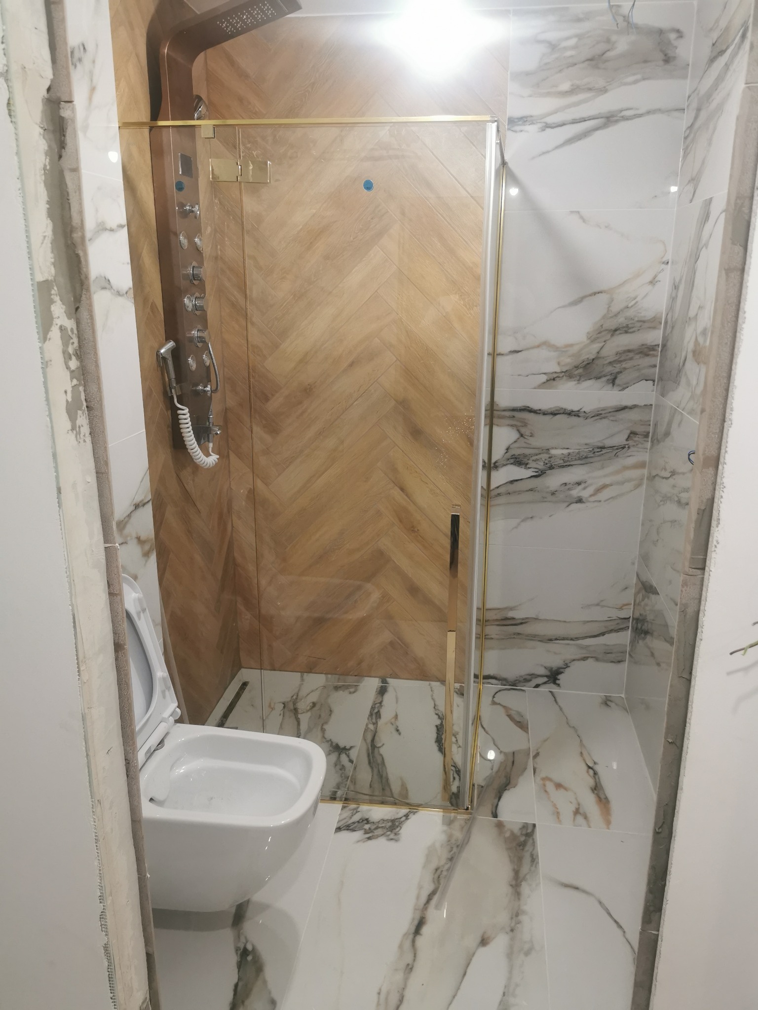 Nowoczesna łazienka z kabiną prysznicową ze złotymi detalami, drewnopodobną ścianą w jodełkę wewnątrz kabiny i marmurowymi płytkami na podłodze i ścianach. W tle widoczna podwieszana miska WC.