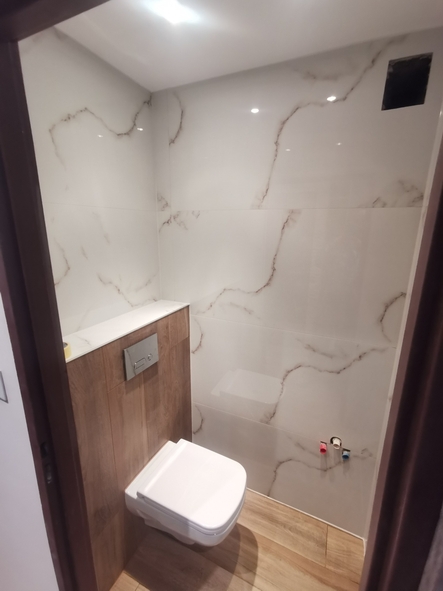 Nowoczesna, mała łazienka po remoncie: podwieszana toaleta z chromowanym przyciskiem spłukiwania, ściany wyłożone białymi płytkami imitującymi marmur z brązowymi żyłkami, drewnopodobne panele...