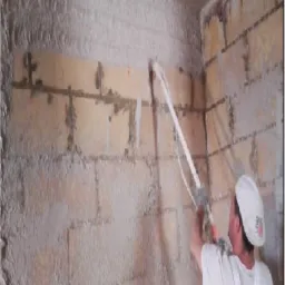 Pracownik w białym kombinezonie aplikuje maszynowo mokrą zaprawę na ścianę z bloczków, widoczne ślady świeżo nałożonego materiału na ścianie i suficie.