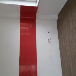 Wnętrze łazienki po remoncie: białe ściany z pionowym pasem czerwonych płytek, ściana z brązowych płytek imitujących drewno, widoczne otwory na instalacje.