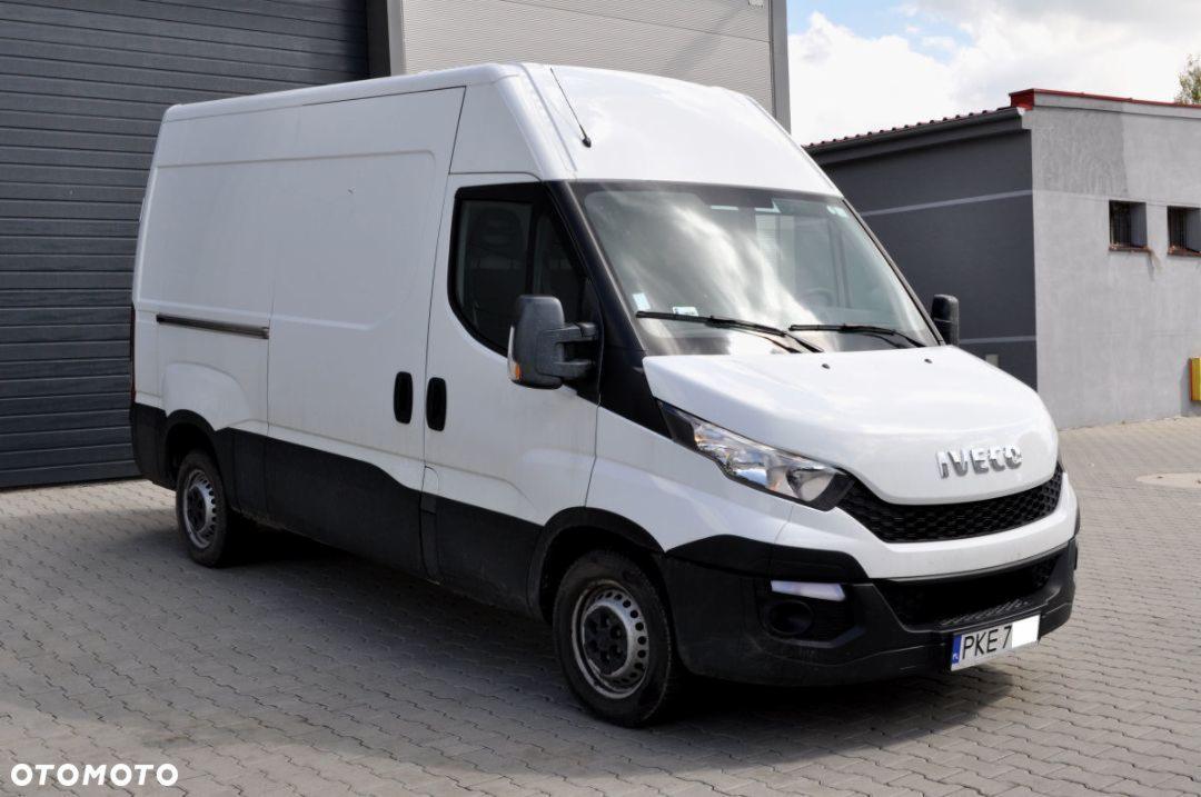 Biały samochód dostawczy Iveco z czarnym dołem zaparkowany na kostce brukowej przed szarą halą, widoczne tablice rejestracyjne PKE7.