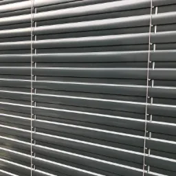 Szare poziome lamele żaluzji, widoczne sznurki sterujące, oświetlone naturalnym światłem.