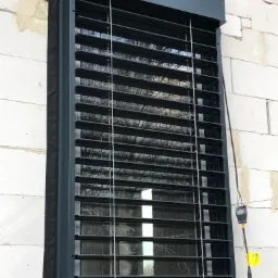 Zamontowana, grafitowa osłona okienna z lamelami na budynku z białych bloczków, widoczne przewody elektryczne.