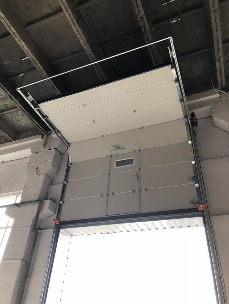 Otwarta segmentowa brama garażowa z małym oknem i drzwiami pośrodku, widok od dołu na betonowy sufit magazynu.