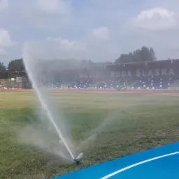 Automatyczny system nawadniania trawy na stadionie MOSiR Ruda Śląska, zraszacz w akcji, w tle trybuny i zieleń