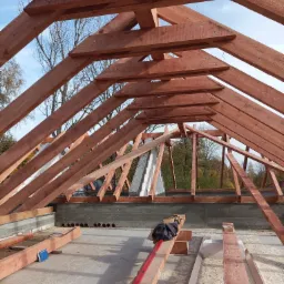 Wnętrze konstrukcji dachu z belek drewnianych w kolorze czerwono-brązowym, widok na niebieskie niebo i drzewa w tle, poziomica i notatnik na betonowej powierzchni.