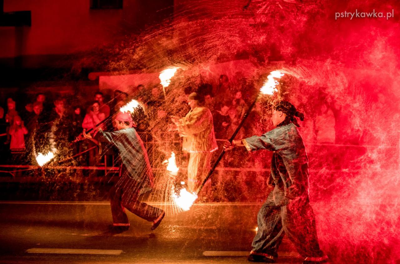 Uliczny pokaz z ogniem: trójka performerów w kostiumach tańczy z płonącymi pochodniami, otoczona fontanną iskier na tle obserwującego tłumu.