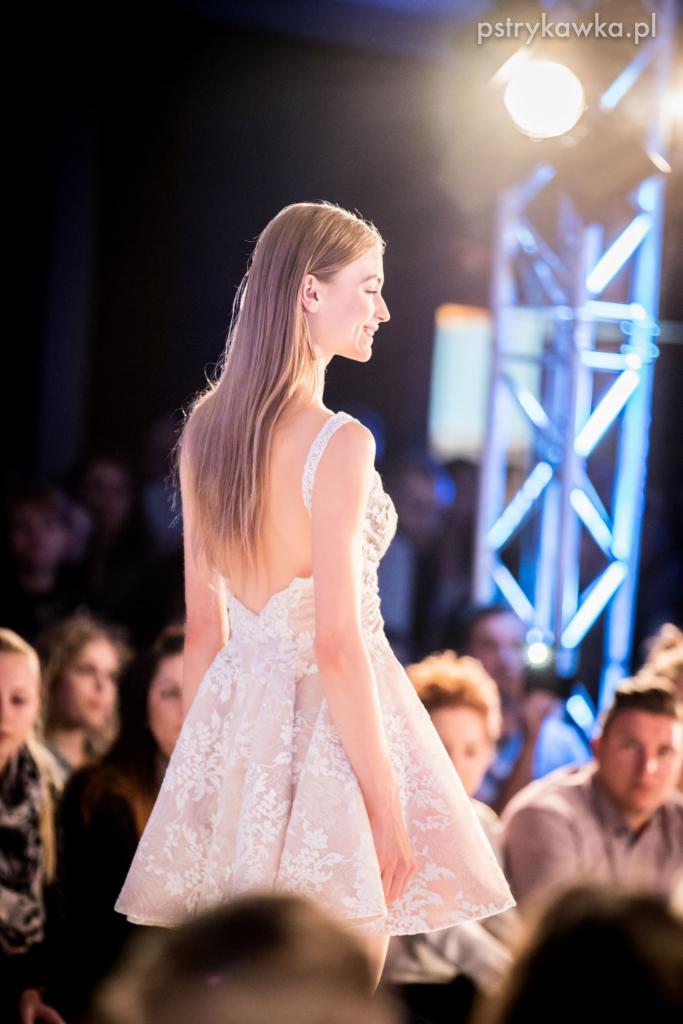 Modelka prezentuje krótką, koronkową sukienkę z odkrytymi plecami na tle sceny z oświetleniem i widownią.