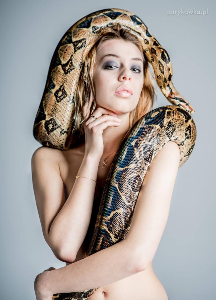 Portret kobiety z wężem boa dusicielem owiniętym wokół szyi i ramion, subtelny makijaż, minimalistyczna biżuteria, gładkie tło.