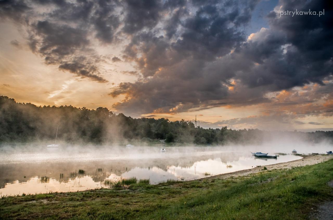 Poranny krajobraz jeziora w Sosnowcu spowitego mgłą, z łodziami na wodzie i dramatycznym niebem o wschodzie słońca.