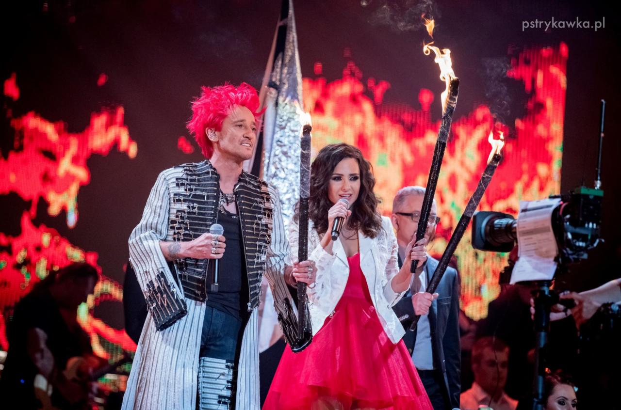 Dwoje artystów na scenie: mężczyzna z różowymi włosami w ekstrawaganckiej marynarce i kobieta w czerwonej sukience trzymający płonące pochodnie, mikrofony w dłoniach, w tle rozświetlona scena.