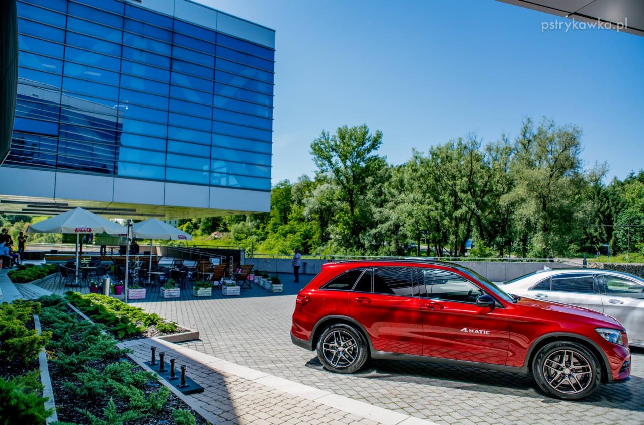 Czerwony samochód typu SUV zaparkowany przed nowoczesnym budynkiem biurowym z przeszkloną fasadą, w tle widoczna zieleń drzew i parasole kawiarniane.