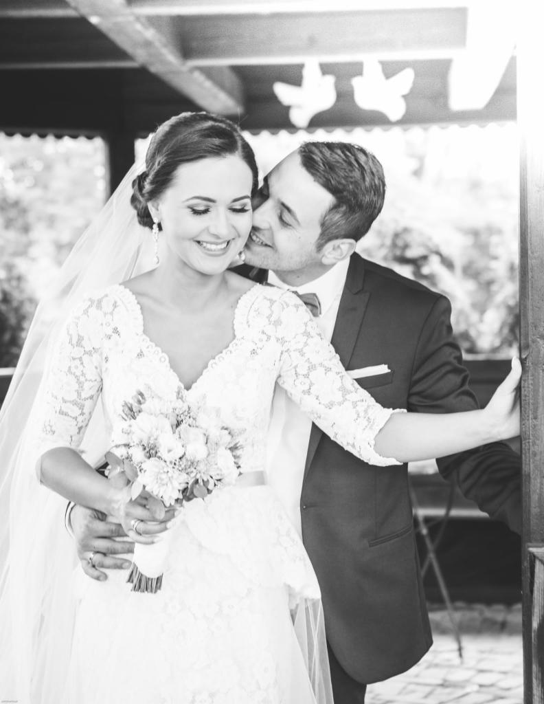 Czarno-białe zdjęcie uśmiechniętej panny młodej w koronkowej sukni z bukietem, obejmowanej przez pana młodego w garniturze, który delikatnie całuje ją w policzek na tle drewnianej konstrukcji.