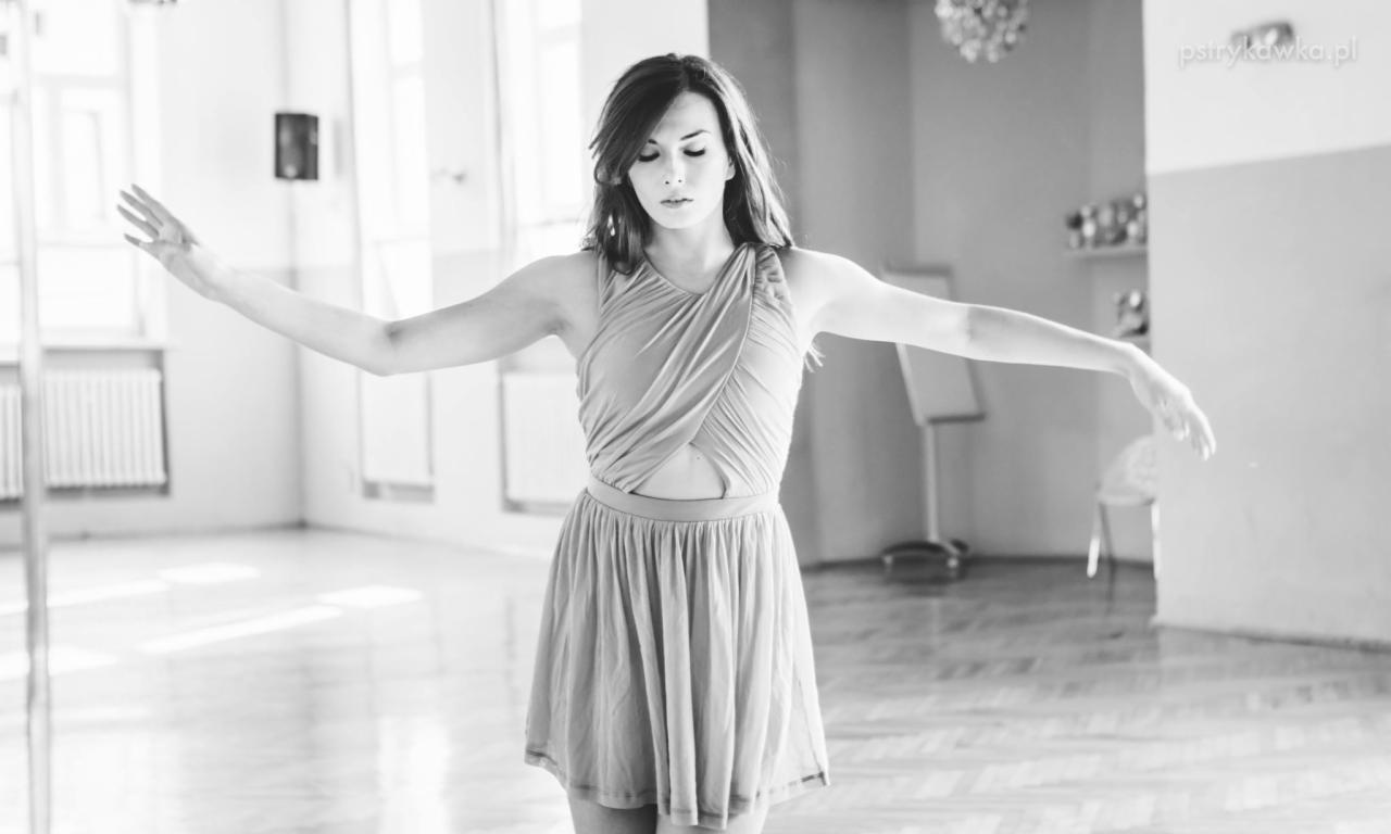 Czarno-białe zdjęcie kobiety w zwiewnej sukience, z rozłożonymi ramionami, w tanecznej pozie w jasnym wnętrzu z parkietem.