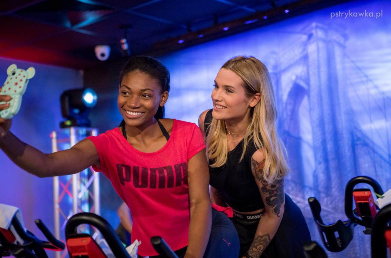 Dwie uśmiechnięte kobiety, jedna robiąca selfie na rowerach stacjonarnych w klubie fitness z tłem przedstawiającym most.