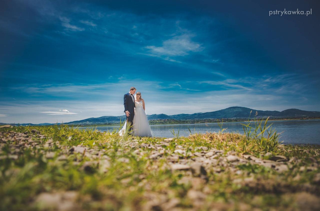Para młoda w strojach ślubnych stoi na tle jeziora i gór, uchwycona z niskiej perspektywy z widocznymi kamieniami i trawą na pierwszym planie, pod błękitnym niebem z lekkimi chmurami.
