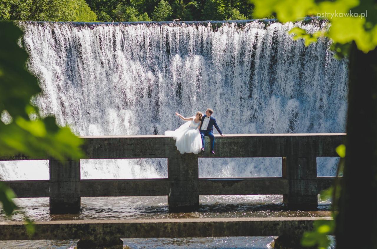 Para młoda siedząca na betonowym murku z wodospadem w tle, panna młoda w białej sukni z odkrytymi ramionami, pan młody w granatowym garniturze, zielone drzewa w tle i w kadrze.