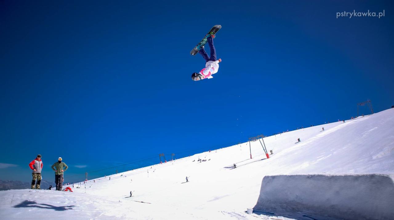 Snowboardzista wykonujący trik w powietrzu na tle błękitnego nieba, widok z dołu stoku, na którym widać wyciągi i narciarzy.