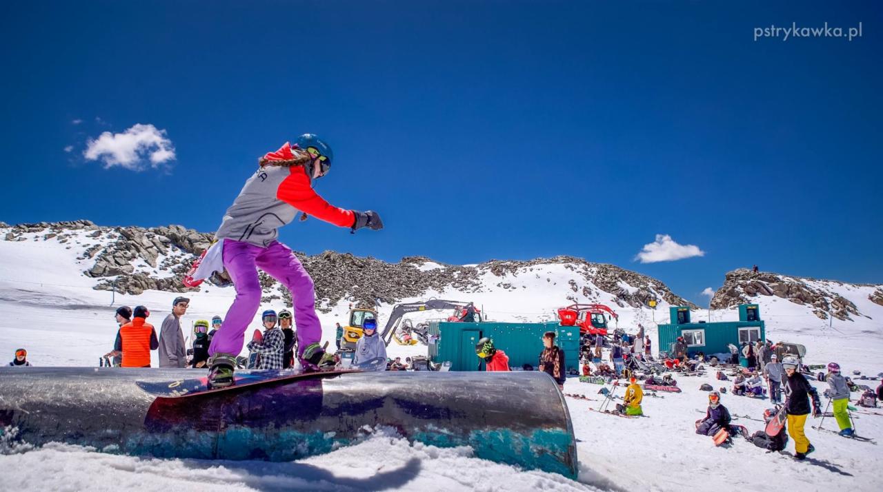 Snowboardzistka w kasku i fioletowych spodniach wykonuje trik na przeszkodzie w snowparku, w tle góry, błękitne niebo i grupa obserwujących osób.