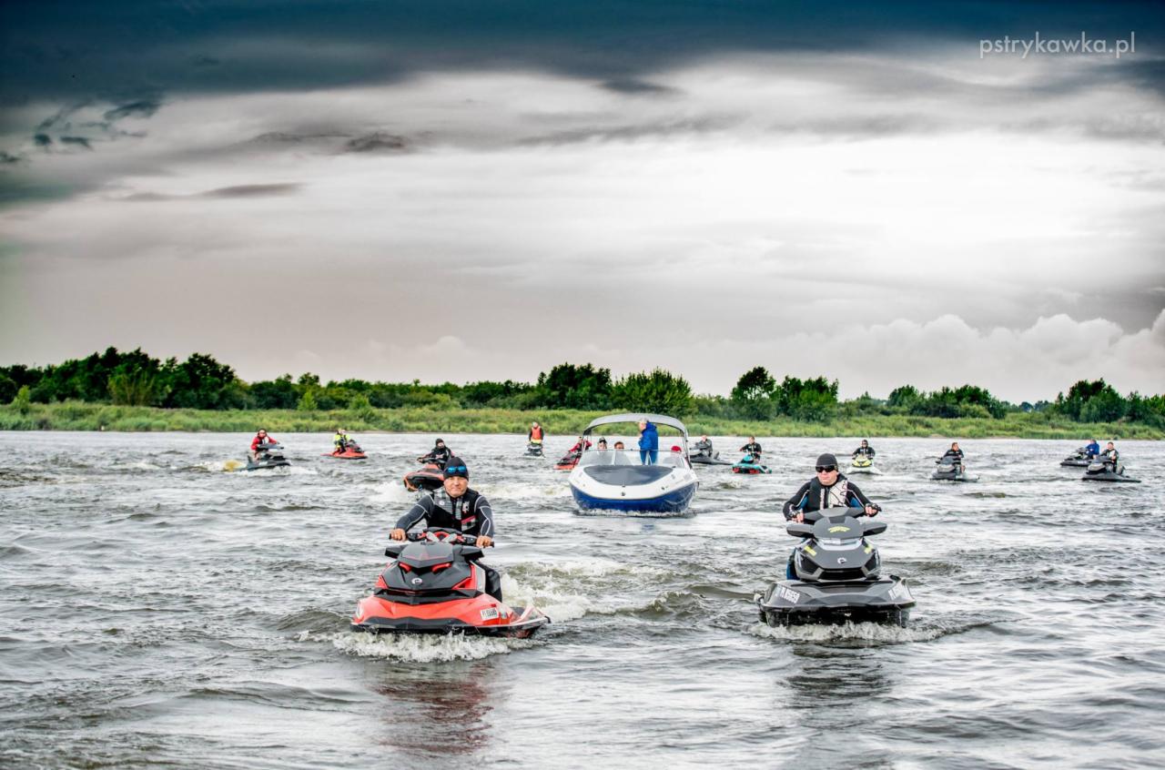 Grupa osób płynie skuterami wodnymi po rzece, w tle łódź motorowa i pochmurne niebo.