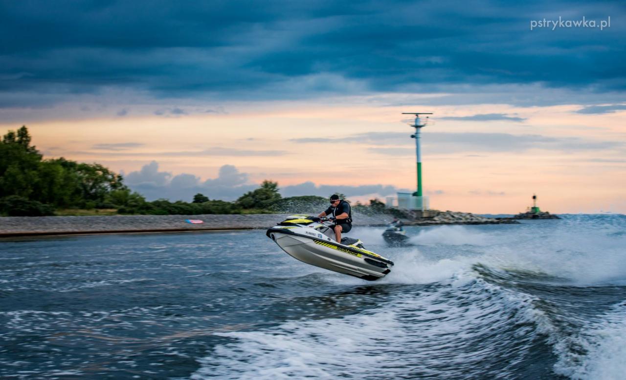 Mężczyzna skacze na skuterze wodnym Sea-Doo z żółtymi akcentami na tle zachmurzonego nieba i portu z latarnią morską.