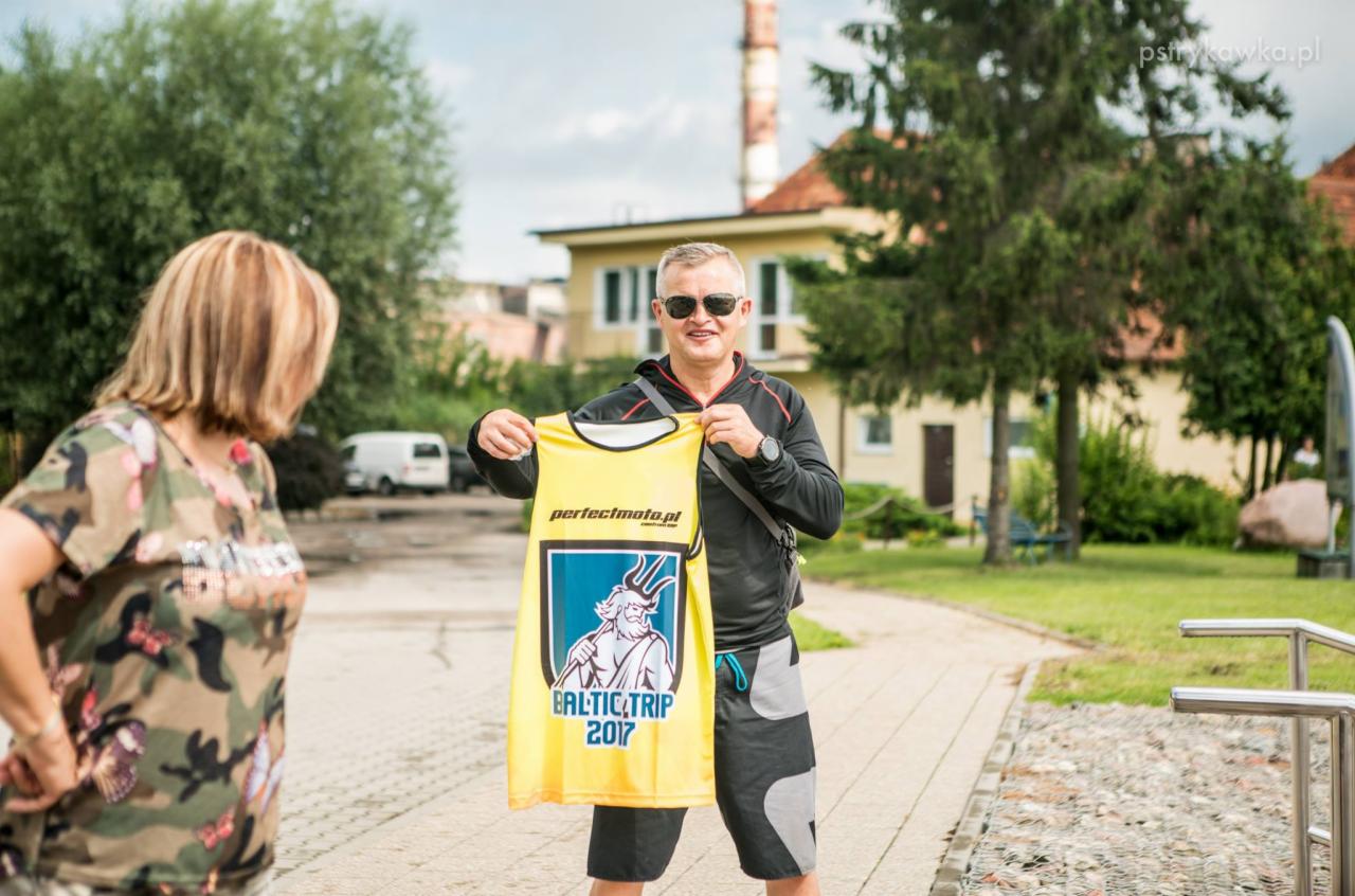 Mężczyzna w okularach słonecznych prezentuje żółtą koszulkę z nadrukiem 'BALTIC TRIP 2017' na tle budynku i drzew, z kobietą w kamuflażowej koszulce w kadrze.