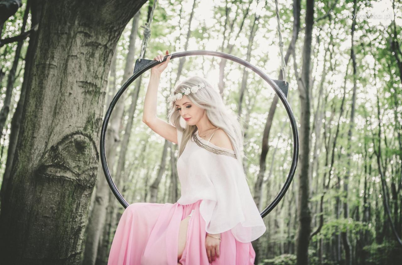 Kobieta z wiankiem na głowie, w zwiewnej białej bluzce i długiej różowej spódnicy z rozcięciem, siedzi na metalowym okręgu zawieszonym w lesie, trzymając się go jedną ręką.