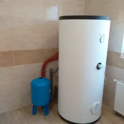 Bojler elektryczny marki Kospel z manometrem i podłączonym przewodem zasilającym, obok niebieskie naczynie przeponowe, całość zainstalowana w pomieszczeniu wyłożonym płytkami.