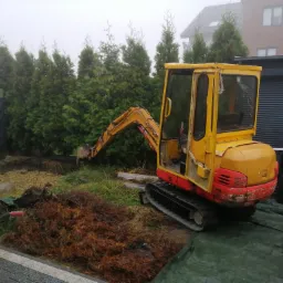 Żółto-czerwona mini koparka podczas prac ziemnych w Knurowie, widoczny pracownik z taczką i pryzma wykopanej ziemi, w tle rząd tuj i mglista pogoda.