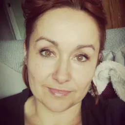 Portret kobiety o rudych włosach, z delikatnym uśmiechem, ujęcie z bliska, w tle drewniana ściana i fragment szarej tkaniny.