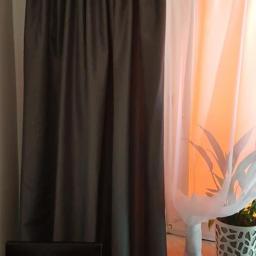 PIĄTKA bis - Zasłona zaciemniająca w kolorze grafitowym, z dekoracyjną falbaną u góry, obok firanka z białego woalu, w tle pomarańczowe światło i roślina doniczkowa na parapecie, fragment krzesła z czerwonym...