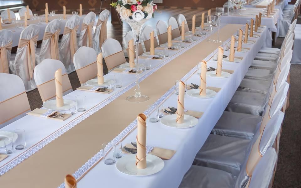 Uroczysty stół weselny z białym obrusem, beżowym bieżnikiem z koronką, elegancko złożonymi serwetkami, szklankami i sztućcami, krzesła z białymi pokrowcami i beżowymi szarfami, centralny wazon...