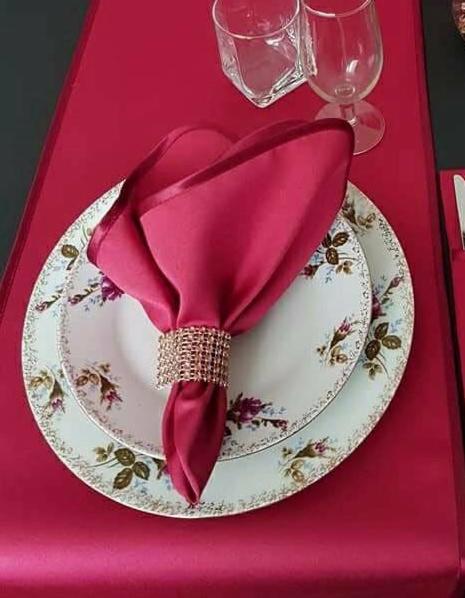 Elegancko nakryty stół z bordowym bieżnikiem, talerz z kwiatowym wzorem, bordowa serwetka ze zdobionym pierścieniem, kieliszki i sztućce.