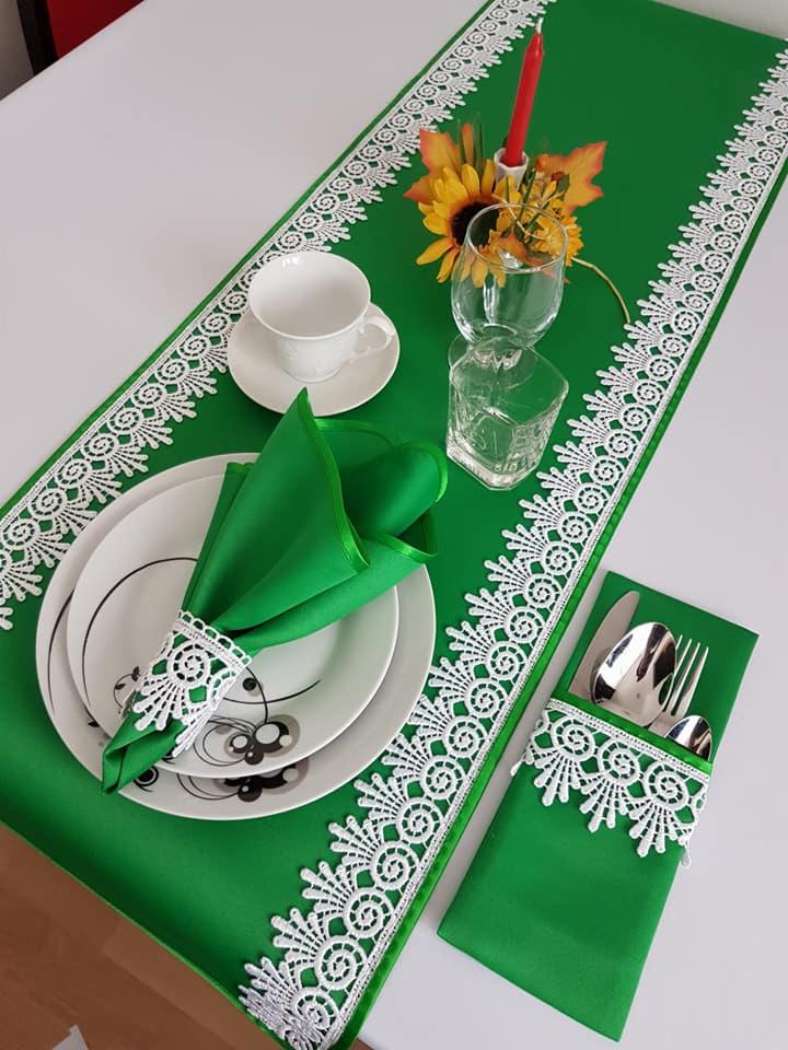 Uroczysty stół nakryty zielonym bieżnikiem z białą koronką, pasujące serwetki z koronkowymi pierścieniami, zastawa stołowa i dekoracje kwiatowe.