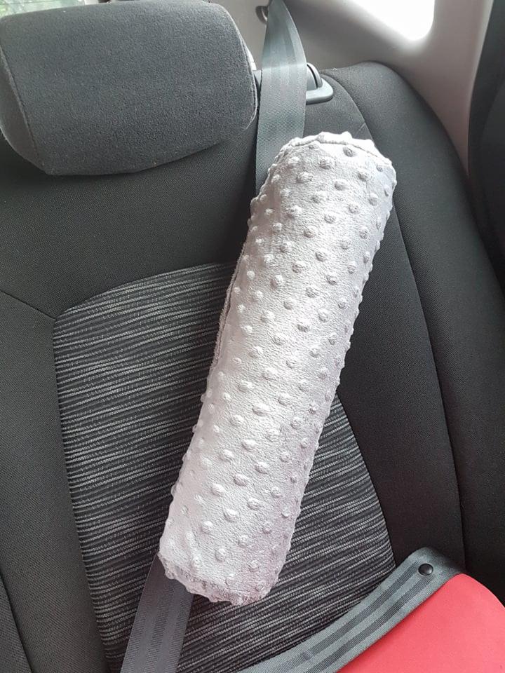 Szary, pluszowy ochraniacz na pas bezpieczeństwa w samochodzie, z wytłaczanymi kropkami, założony na pas w czarnym fotelu z szarymi paskami.