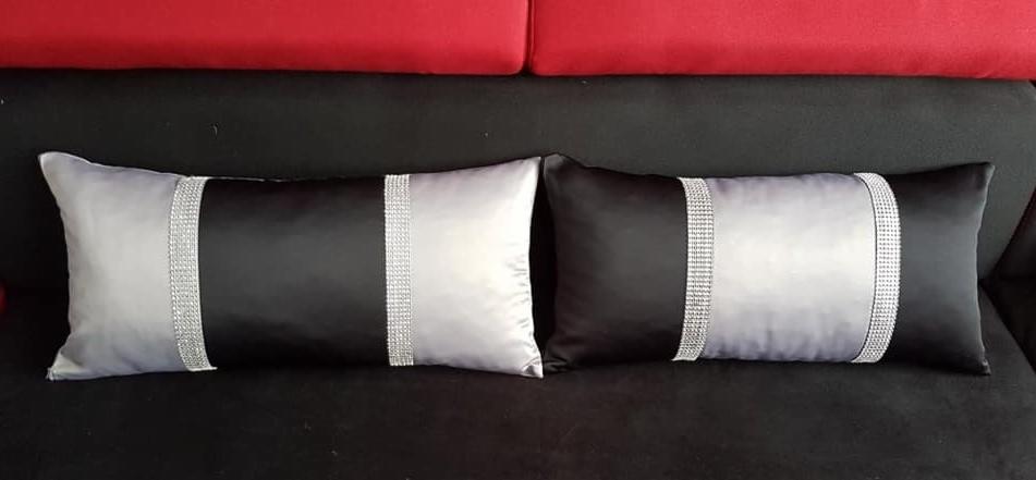 Dwie dekoracyjne poduszki w kolorach czerni i srebra z pasem cyrkonii, leżące na czarnej sofie z czerwonym zagłówkiem w tle.