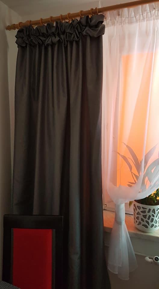 Zasłona zaciemniająca w kolorze grafitowym, z dekoracyjną falbaną u góry, obok firanka z białego woalu, w tle pomarańczowe światło i roślina doniczkowa na parapecie, fragment krzesła z czerwonym...