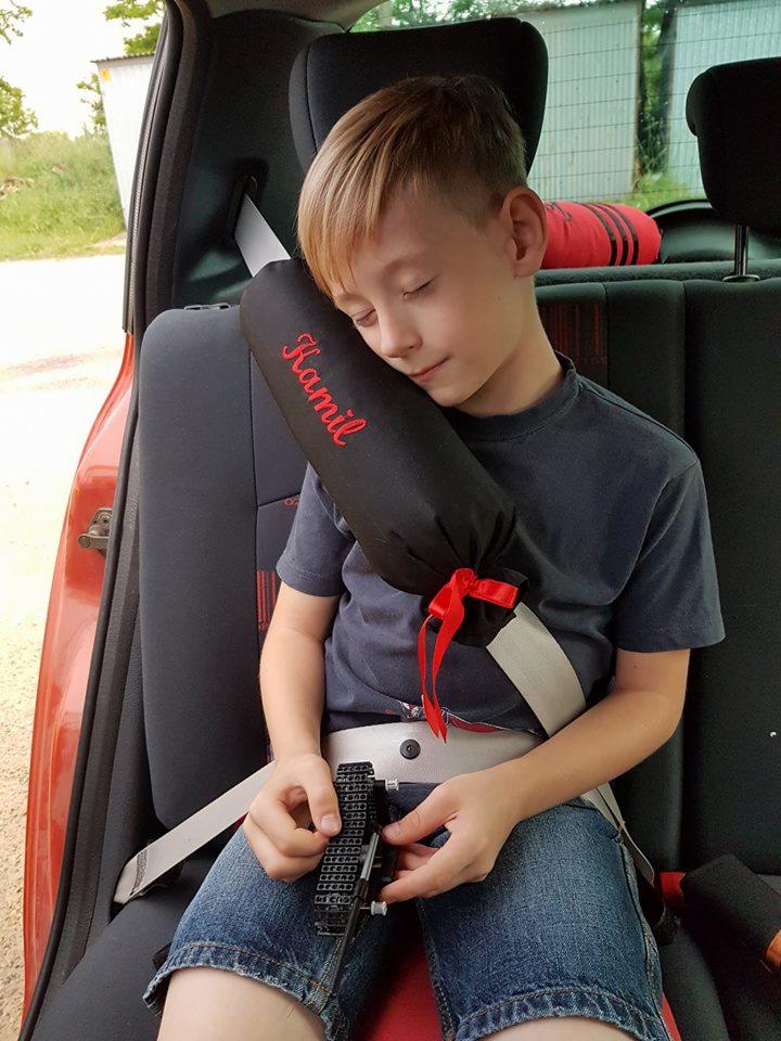 Chłopiec śpi w samochodzie, opierając głowę o ochraniacz na pas z wyhaftowanym imieniem 'Kamil', trzymając w rękach zabawkę.