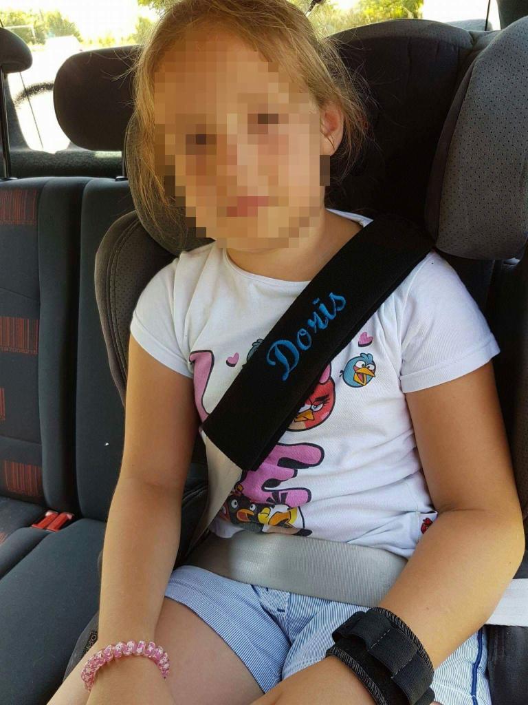 Dziewczynka w foteliku samochodowym z czarnym ochraniaczem na pas z wyhaftowanym imieniem 'Doris', ubrana w białą koszulkę z nadrukiem Angry Birds.