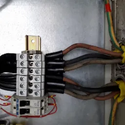 Szczegółowe ujęcie rozdzielnicy elektrycznej z widocznymi przewodami, listwą zaciskową i zabezpieczeniami, zamontowanej w metalowej obudowie.