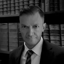 Portret mężczyzny w garniturze i krawacie na tle regału z książkami, zdjęcie czarno-białe, ostre światło z góry.