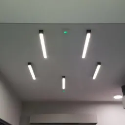 Nowoczesne oświetlenie sufitowe z pięcioma podłużnymi lampami LED o czarnych obudowach, rozmieszczonymi symetrycznie na szarym suficie, z dodatkowym czujnikiem z zieloną diodą.