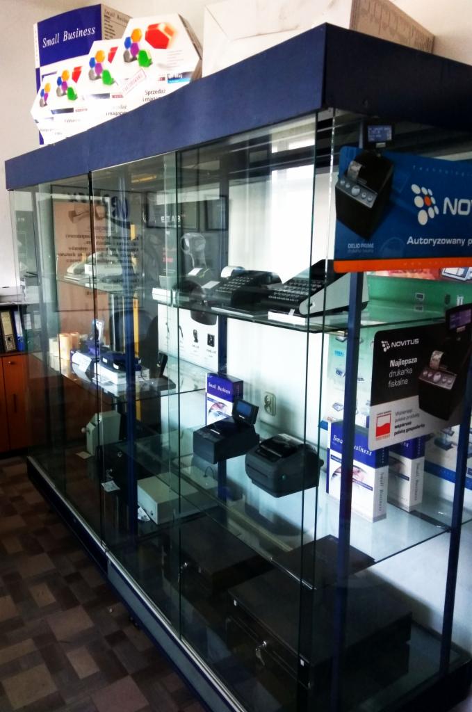 Szklana witryna sklepowa z kasami fiskalnymi, drukarkami fiskalnymi i akcesoriami biurowymi, w tle ściana i szafka.