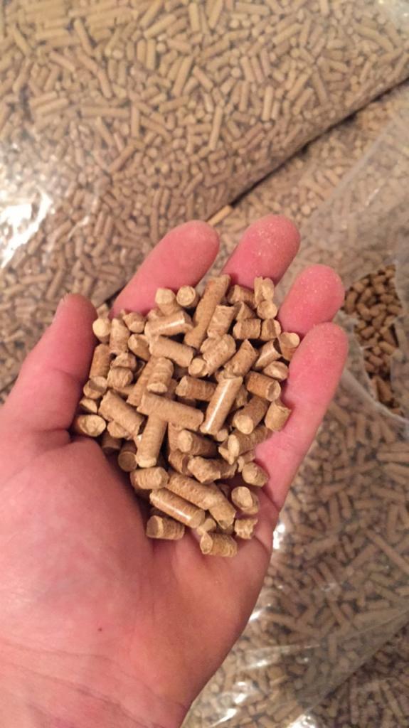 Otwarta dłoń trzymająca garść jasnych, drewnianych peletek na tle worków wypełnionych tym samym materiałem.