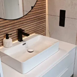 Nowoczesna łazienka z białą, prostokątną umywalką nablatową, czarną armaturą i okrągłym lustrem z drewnianym akcentem, na tle beżowych płytek.